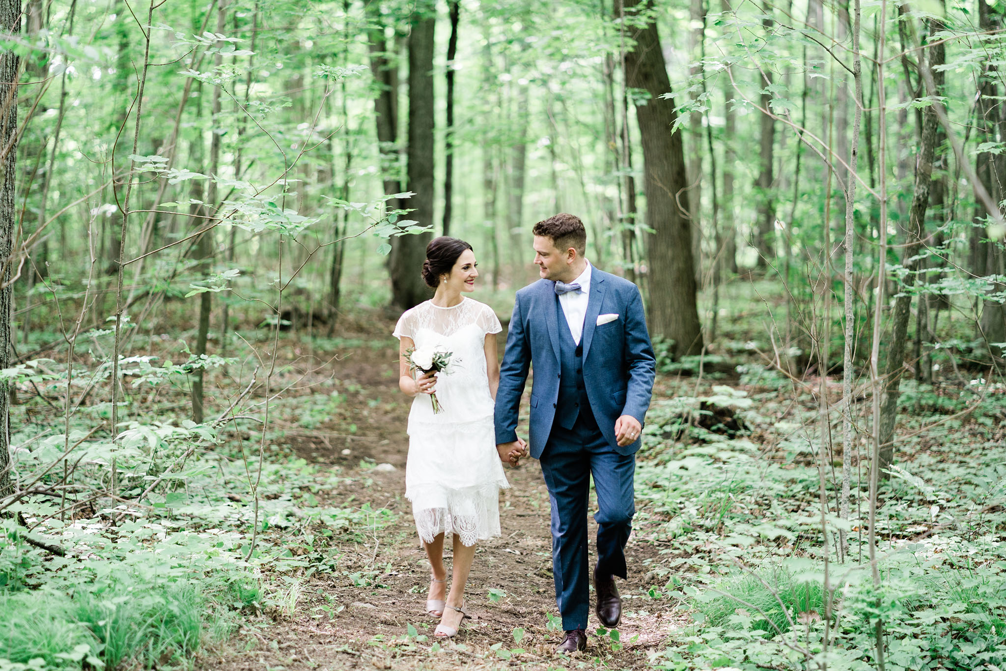Happily married couple waking thru a quiet and peaceful forest hand in hand, just after getting married. Bride wearing cocktail length dress reminiscent of a 1930's flapper. Gentleman wearing dark blue suit with bowtie. Floral arrangement is small, delicate and classic.