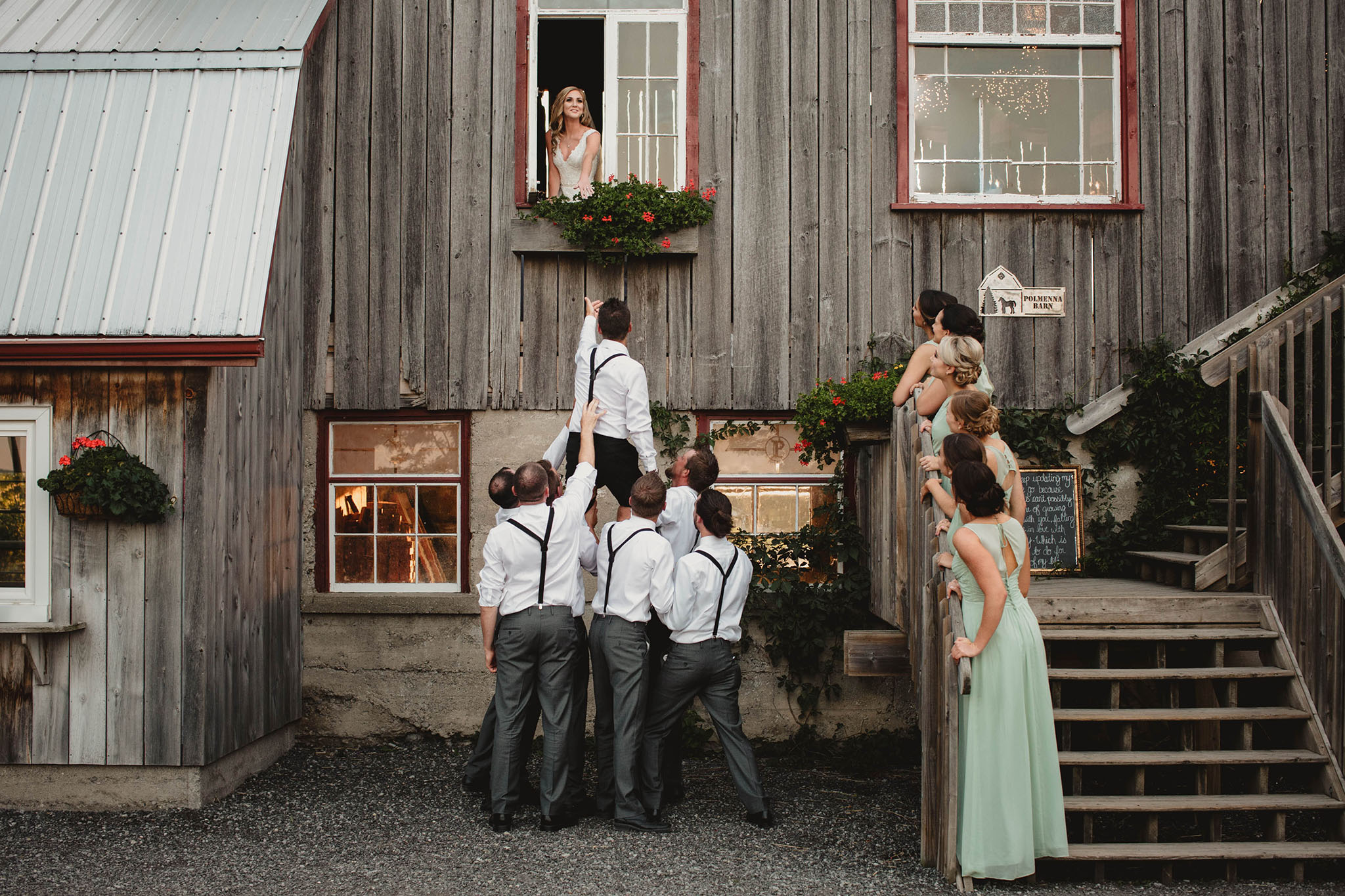 Bride looking out Barn window and below are the groom with the groomsmen lifting him up to the window.