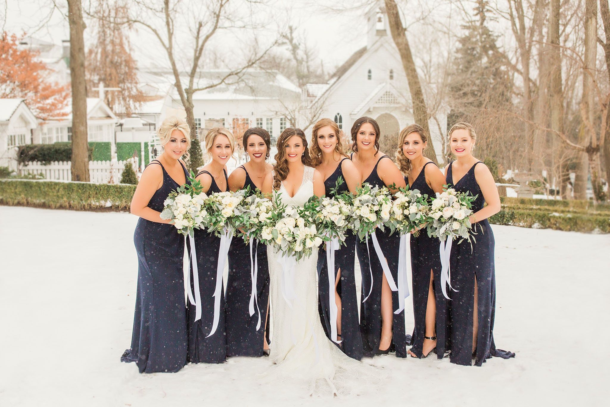 Eight beautiful ladies all outside with snow all around. Standing in front of heritage house with green hedges and gorgeous white floral bouquets.
